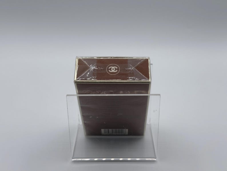 May include: A brown rectangular box with a gold trim and a clear plastic wrap. The top of the box features a white logo. The box is displayed on a clear acrylic stand.
