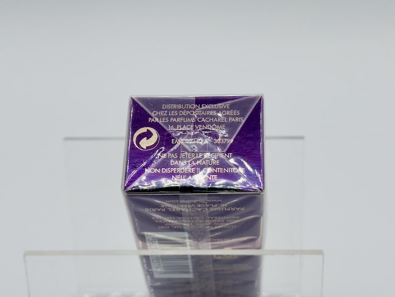 May include: A purple perfume box with white text and a recycling symbol. The text includes the words "DISTRIBUTION EXCLUSIVE" and "PARFUMS CACHAREL PARIS". The box is on a clear shelf.