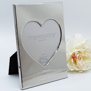 May include: Silver heart-shaped photo frame with a 5x5 inch opening. The frame has a textured surface and is engraved with the text "Impressions Photo Frame", "5x5 in 13x13cm approx", "Lacquer Coated Tarnish Resist", "Susan 25 Years of happiness 1999-2024".