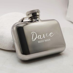 May include: A polished silver hip flask with a textured screw cap and hinged lid. The flask is engraved with the name "Dave" and the words "BEST MAN" in white script. The flask has a rectangular shape.