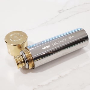 May include: A silver and gold flask with a gold cap. The flask is engraved with the text "CARL | HAPPY 50TH" and a small moustache.