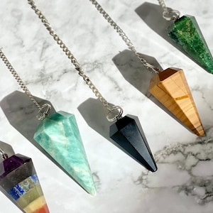 Op de afbeelding: Een verzameling kristallen pendels met zilveren kettingen. De pendels zijn in verschillende kleuren, waaronder groen, zwart en een regenboog aan kleuren. De pendels worden gepresenteerd op een marmeren oppervlak.