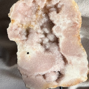 May include: A pink and white geode with a large opening revealing a cluster of white crystals. The geode is mounted on a black metal stand.