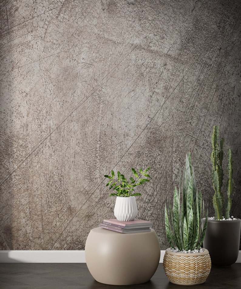 May include: A round, light brown side table with a stack of books and a white ceramic vase with a green plant.  There are two potted plants on the floor, one with a woven basket and the other in a black pot. The background is a textured, gray wall.
