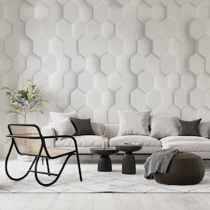 May include: A white couch with three pillows and a brown ottoman in front of a white wall with a hexagonal pattern. There are two black side tables with a vase on one of them.