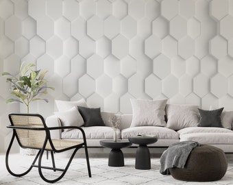 May include: A white couch with three pillows and a brown ottoman in front of a white wall with a hexagonal pattern. There are two black side tables with a vase on one of them.