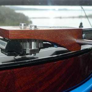 Puede incluir: Primer plano de un brazo de tono de madera en un tocadiscos. El brazo de tono está unido a un brazo de metal plateado con un cartucho negro y una aguja. El tocadiscos tiene un plato negro y una base de madera marrón.