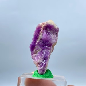 May include: A raw, natural amethyst crystal specimen. The purple crystal has a rough texture and is mounted on a clear acrylic base with a green support. The crystal's color ranges from deep violet to pale lavender.
