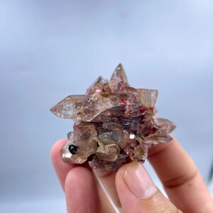 May include: A cluster of natural quartz crystals with reddish-brown and clear hues. The crystals are arranged in a layered formation, with sharp, pointed tips. A small, dark inclusion is visible on the front. The overall shape resembles a small animal.