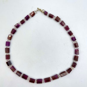 Può includere: Una collana di pietre preziose viola e trasparenti con una chiusura dorata. La collana è composta da perline quadrate.