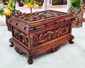 May include: A dark brown wooden box with intricate carvings and a hinged lid. The box has a decorative design on the front and sides, and it sits on four small feet.