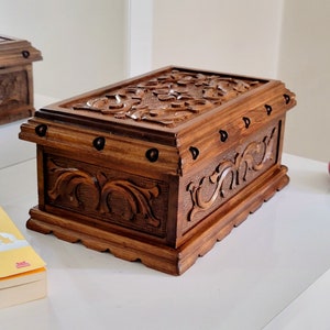 Peut inclure: Une boîte en bois avec des sculptures complexes et des accents métalliques. La boîte est brun foncé et de forme rectangulaire.