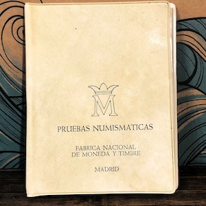 Peut inclure: Un livre de couleur crème avec le titre "PRUEBAS NUMISMATICAS" et le texte "FABRICA NACIONAL DE MONEDA Y TIMBRE MADRID" imprimé sur la couverture.