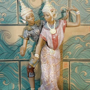 May include: A ceramic sculpture depicting two figures in traditional dance poses. The figures wear ornate, pointed headdresses, necklaces, and bracelets. One figure wears a dark, textured top and trousers, while the other wears a light pink draped garment.