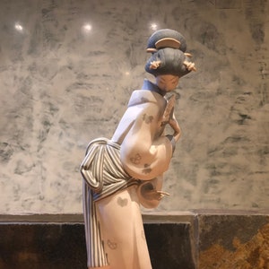 Può includere: Una statuetta in porcellana di una donna in un kimono bianco e blu con un ventaglio in mano. La statuetta è in piedi su una base rotonda con un bordo dorato.