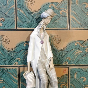 May include: A porcelain figurine of a clown holding an accordion. The clown is wearing a white jacket, plaid trousers, a hat, and has a sad expression. The figurine is standing on a white base.