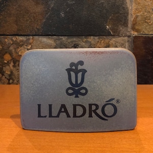 Könnte beinhalten: Eine blaue Keramikfliese mit dem Lladró-Logo, das ein stilisiertes Blumendesign und das Wort "LLADRÓ" in schwarzen Buchstaben zeigt.
