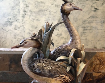 May include: A porcelain figurine of two grebes, one with its head held high and the other with its head turned to the side. The grebes are perched on a nest with three eggs. The figurine is painted in shades of brown, grey, and white.