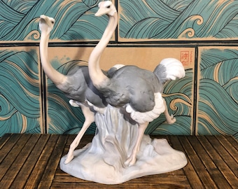 May include: A ceramic sculpture of two ostriches. The birds have grey and white feathers, long necks, and are standing on a white base. The background features a wave pattern in shades of blue and green.
