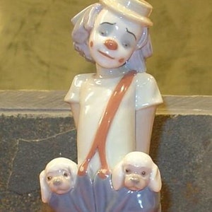 May include: A porcelain figurine of a clown wearing a blue and white outfit with a red and white striped shirt and a red bow tie. The clown is holding two white porcelain puppies in its arms.