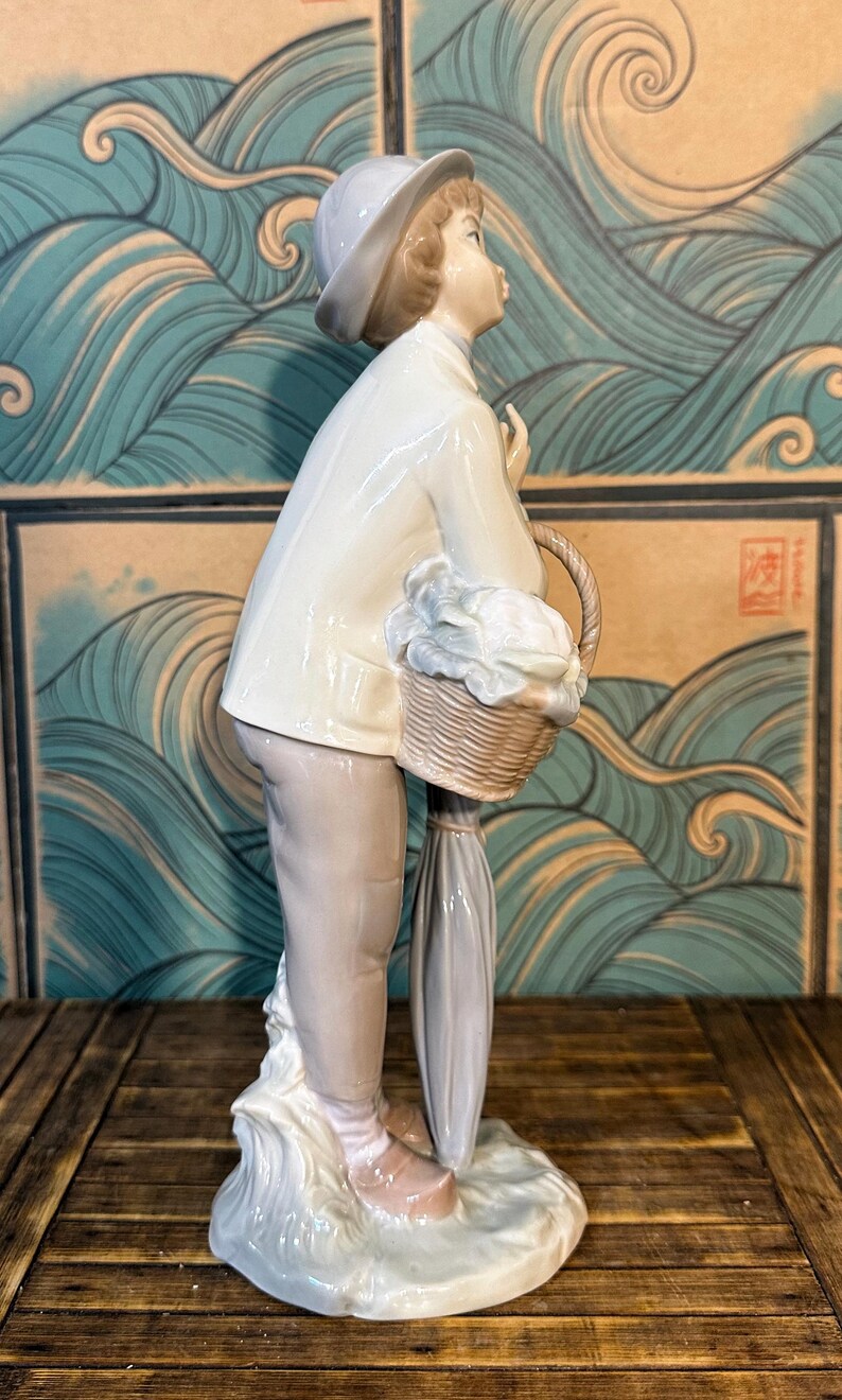 May include: A porcelain figurine of a person wearing a light-coloured jacket, trousers, and a hat. The figure holds a woven basket filled with white items and carries a closed umbrella. The sculpture is finished in a glossy glaze with soft tones.