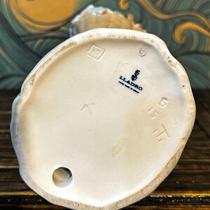 May include: The underside of a white porcelain figurine featuring a blue Lladro logo stamp with the text Hand Made in Spain. The base includes an impressed square mark, a circular hole, and the stamped numbers 5515 on the smooth ceramic surface.