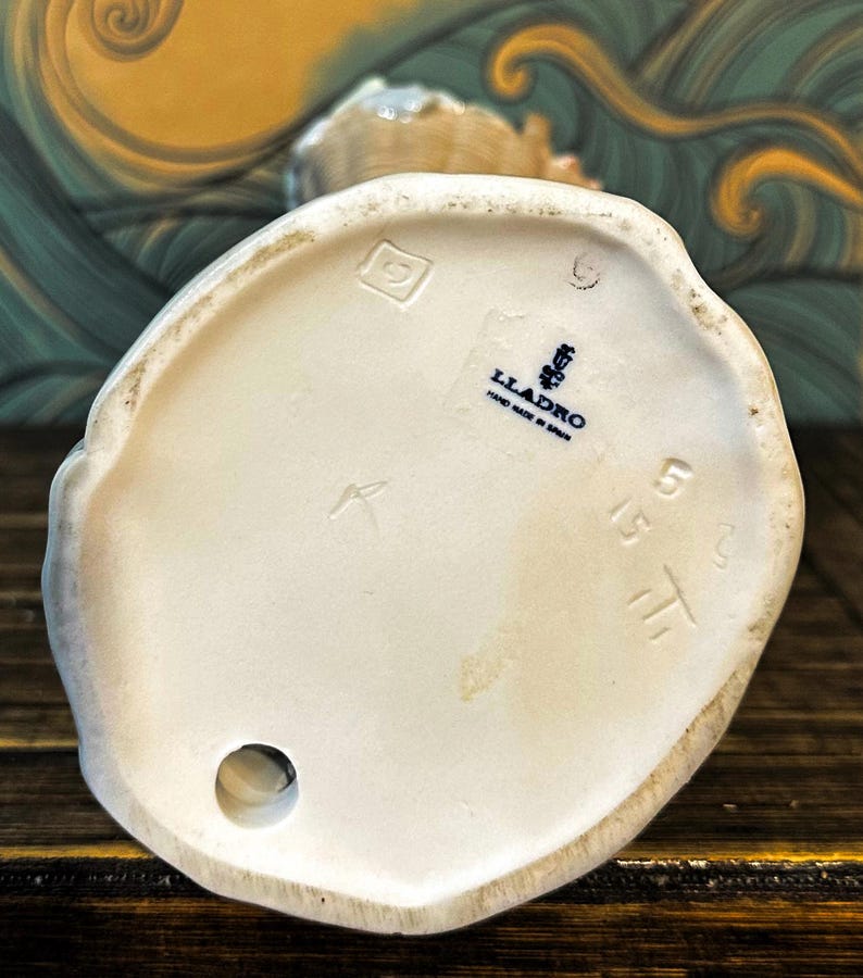 May include: The underside of a white porcelain figurine featuring a blue Lladro logo stamp with the text Hand Made in Spain. The base includes an impressed square mark, a circular hole, and the stamped numbers 5515 on the smooth ceramic surface.