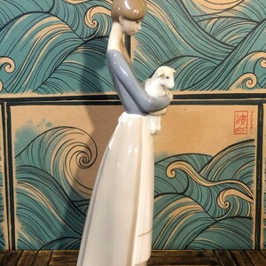 May include: A porcelain figurine of a woman holding a lamb. The woman wears a grey top and a long white dress. The figurine is set against a backdrop of stylized blue waves.