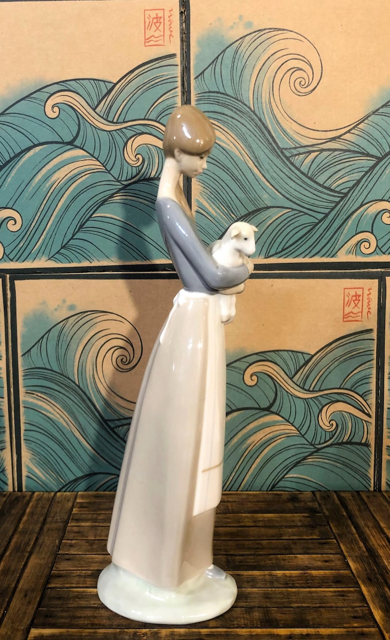 May include: A porcelain figurine of a woman holding a lamb. The woman wears a grey top and a long white dress. The figurine is set against a backdrop of stylized blue waves.