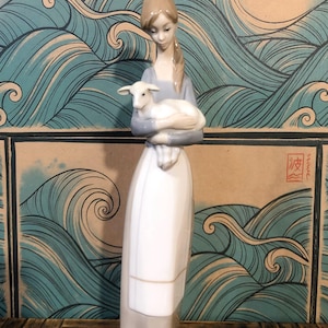 May include: A ceramic figurine depicting a woman cradling a lamb. The woman is dressed in a long white dress and a pale blue top. The backdrop features a design of stylized blue waves.