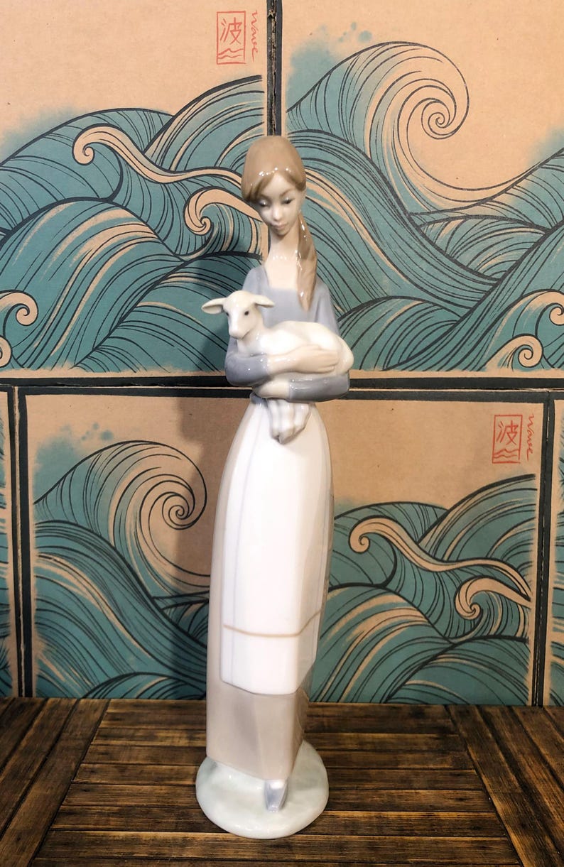 May include: A ceramic figurine depicting a woman cradling a lamb. The woman is dressed in a long white dress and a pale blue top. The backdrop features a design of stylized blue waves.
