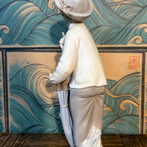 May include: A porcelain figurine of a person wearing a light-coloured hat with a bow, a white collared shirt, and light brown trousers. The figure holds a closed light blue umbrella and stands on a base with sculpted leaf-like details.