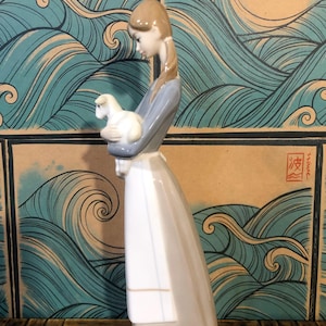 May include: A porcelain figurine of a woman holding a lamb. The woman wears a long, flowing dress in shades of grey and white, with a blue-grey top. The lamb is white. The background features a wave pattern.