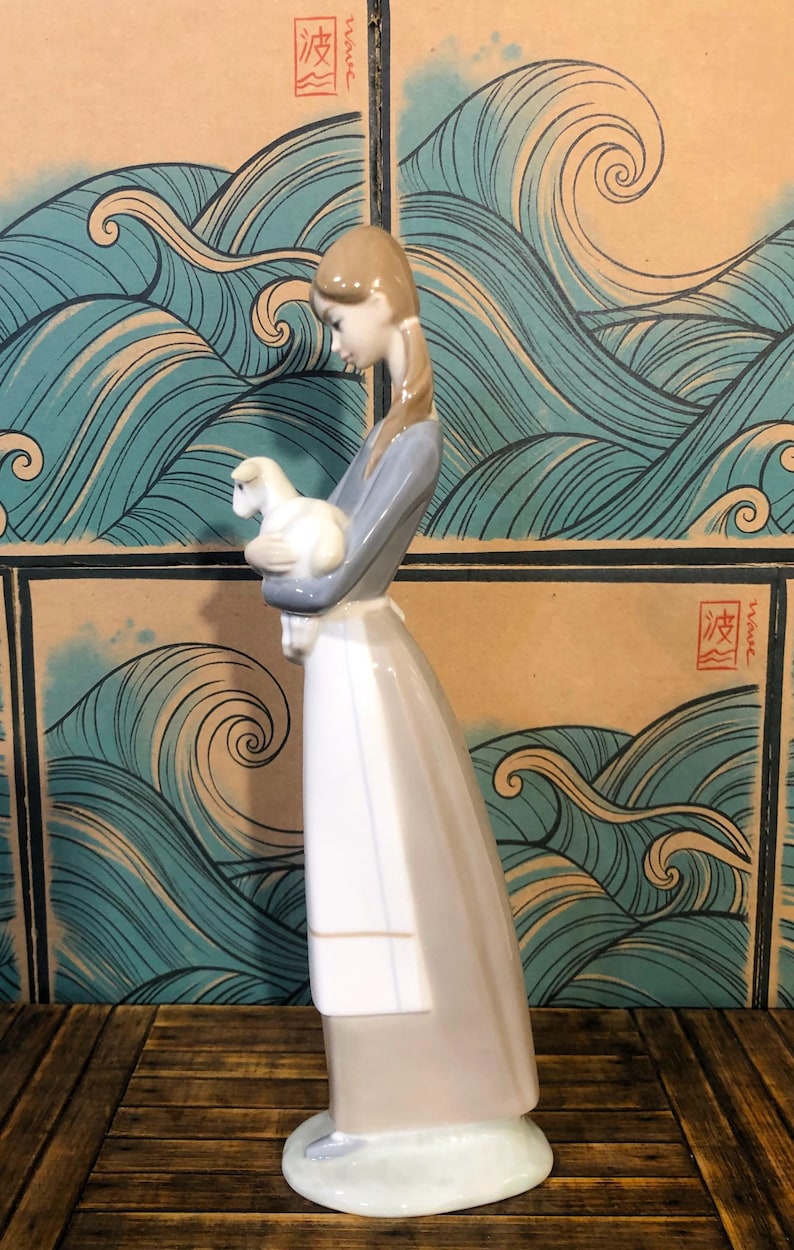 May include: A porcelain figurine of a woman holding a lamb. The woman wears a long, flowing dress in shades of grey and white, with a blue-grey top. The lamb is white. The background features a wave pattern.