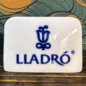 May include: A rectangular white ceramic plaque featuring the blue Lladro logo, which consists of a stylised flower symbol above the brand name text. The surface is smooth and glossy, with rounded corners and a clean, minimalist aesthetic design.