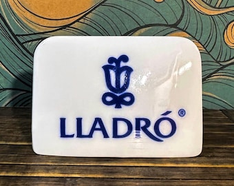 May include: A rectangular white ceramic plaque featuring the blue Lladro logo, which consists of a stylised flower symbol above the brand name text. The surface is smooth and glossy, with rounded corners and a clean, minimalist aesthetic design.