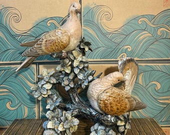 May include: A ceramic figurine featuring two brown and tan doves perched on a dark, textured branch adorned with numerous small, light blue and grey flowers. The birds have detailed feather patterns and are positioned at different heights.