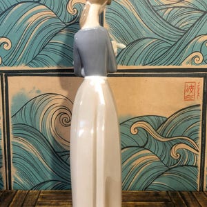 May include: A ceramic figurine of a woman in a long dress, with a grey top and light brown hair. The figure is set against a backdrop of stylised ocean waves in shades of blue and tan.