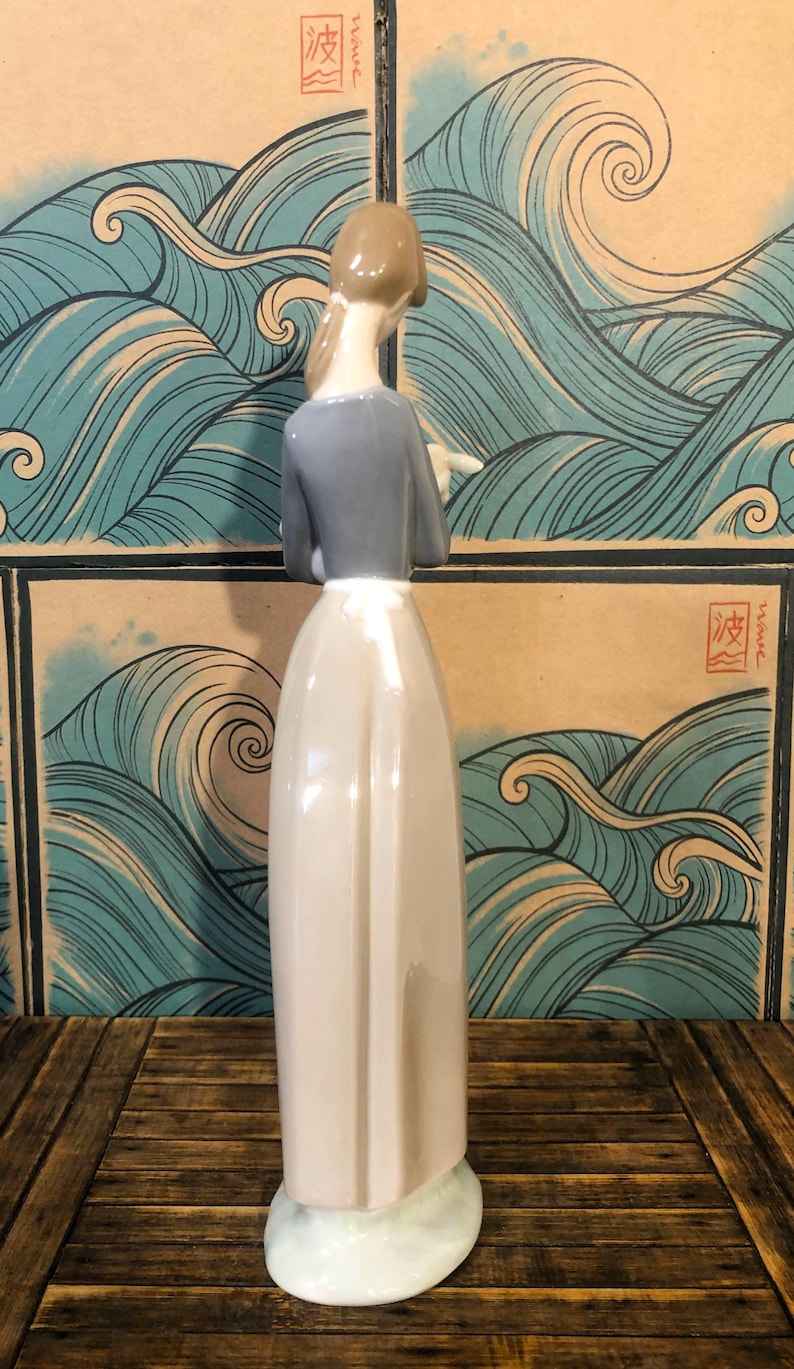 May include: A ceramic figurine of a woman in a long dress, with a grey top and light brown hair. The figure is set against a backdrop of stylised ocean waves in shades of blue and tan.