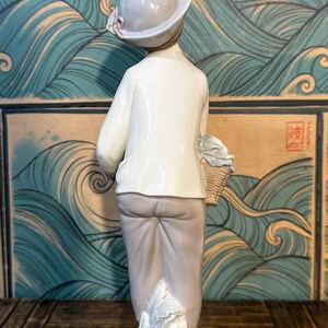 May include: A porcelain figurine of a person wearing a light grey hat, a white long-sleeved shirt, and light grey trousers. The figure holds a small woven basket at their side and stands upon a base sculpted to resemble flowing water or waves.
