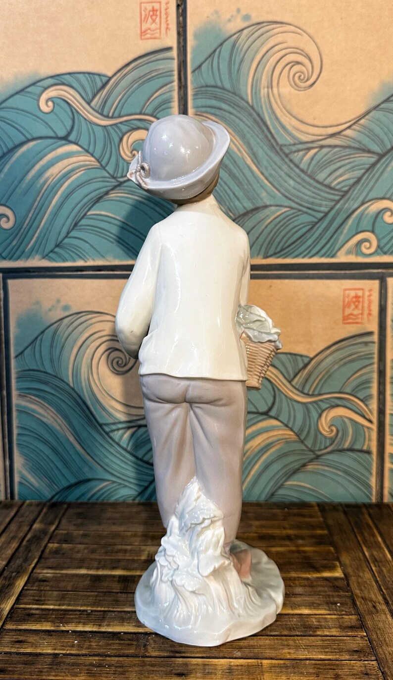 May include: A porcelain figurine of a person wearing a light grey hat, a white long-sleeved shirt, and light grey trousers. The figure holds a small woven basket at their side and stands upon a base sculpted to resemble flowing water or waves.