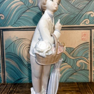 May include: A porcelain figurine of a person wearing a light-coloured jacket, trousers, and a hat. The figure holds a woven basket filled with white and pink items, with a closed umbrella hanging from the arm, standing on a textured base.