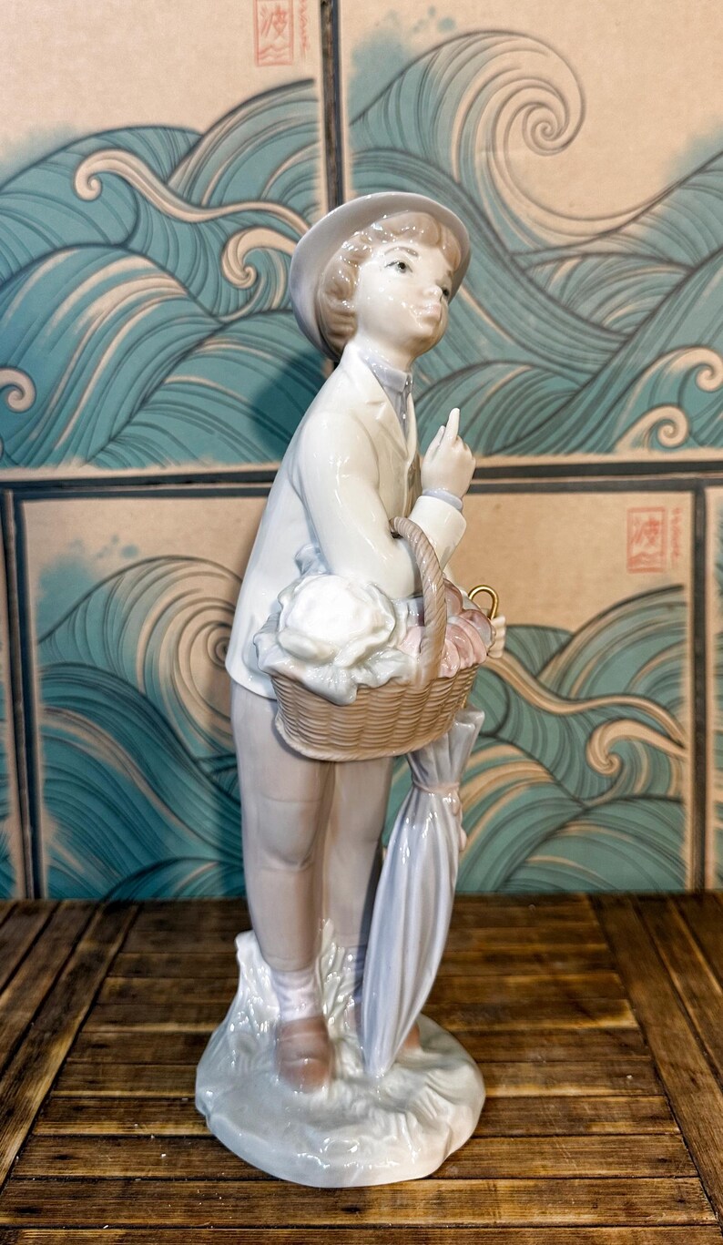 May include: A porcelain figurine of a person wearing a light-coloured jacket, trousers, and a hat. The figure holds a woven basket filled with white and pink items, with a closed umbrella hanging from the arm, standing on a textured base.