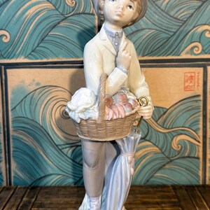 May include: A porcelain figurine of a person wearing a hat, a light-coloured jacket, and trousers, holding a woven basket filled with produce. The figure stands on a base and holds a closed, light blue umbrella in one hand, with a finger raised.