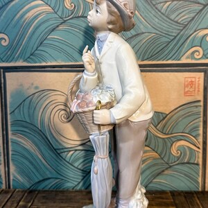 May include: A porcelain figurine of a young person wearing a light-coloured jacket, trousers, and a cap. The figure holds a woven basket filled with items and a closed umbrella with a gold-coloured handle, while looking upward with a finger raised.