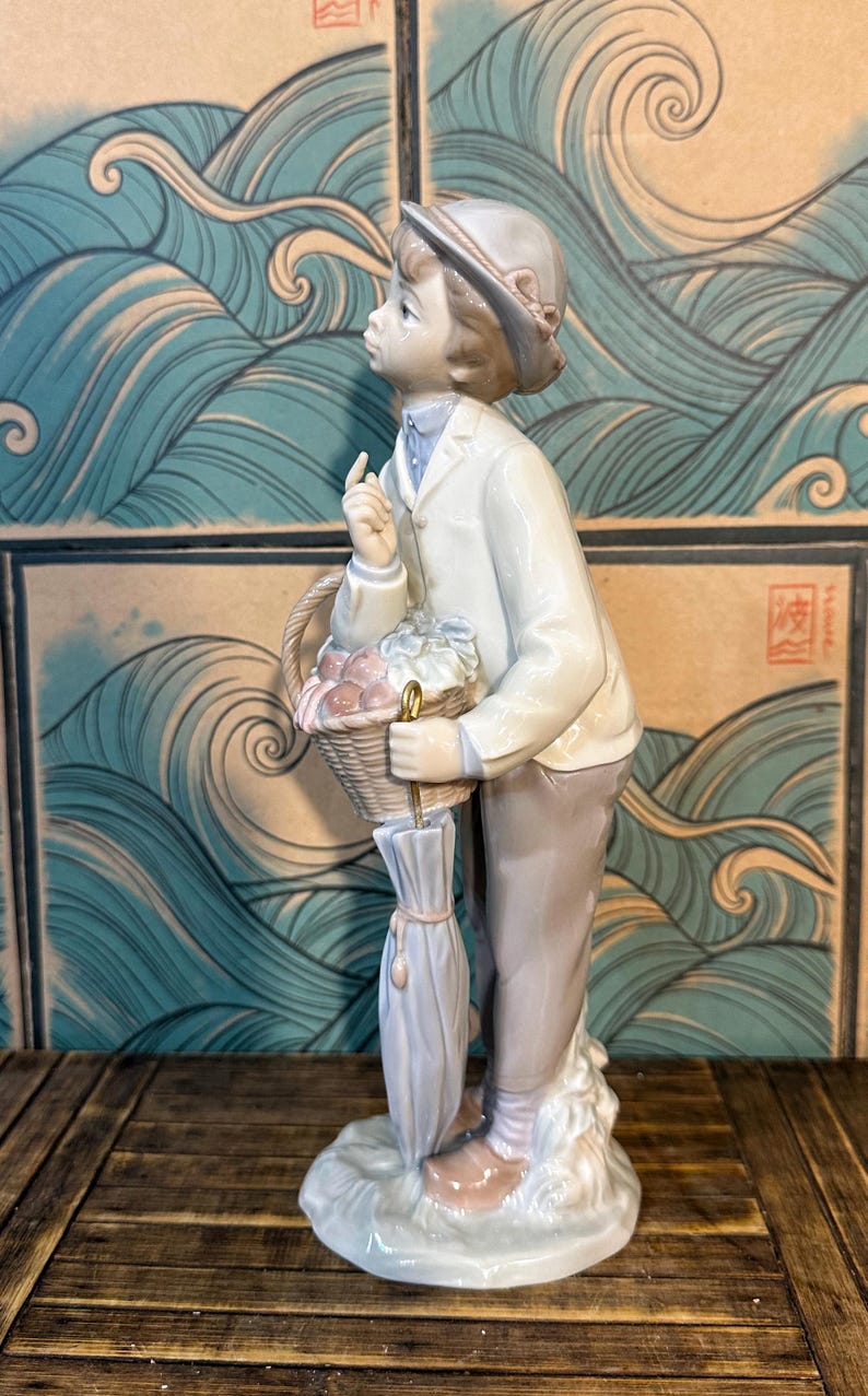 May include: A porcelain figurine of a young person wearing a light-coloured jacket, trousers, and a cap. The figure holds a woven basket filled with items and a closed umbrella with a gold-coloured handle, while looking upward with a finger raised.