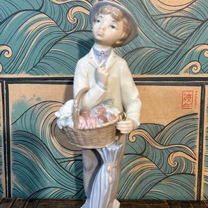 May include: A porcelain figurine of a person wearing a hat and light-coloured clothing, holding a woven basket filled with round objects. The figure stands on a textured base and holds a closed, light blue umbrella in one hand, with a finger raised near the chin.