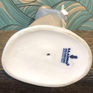 May include: A porcelain figurine of a person with a rabbit. The figurine is primarily white, with accents of blue, tan, and brown. The base is oval-shaped and white, with the Lladro logo and text. The figurine is on a wooden surface.