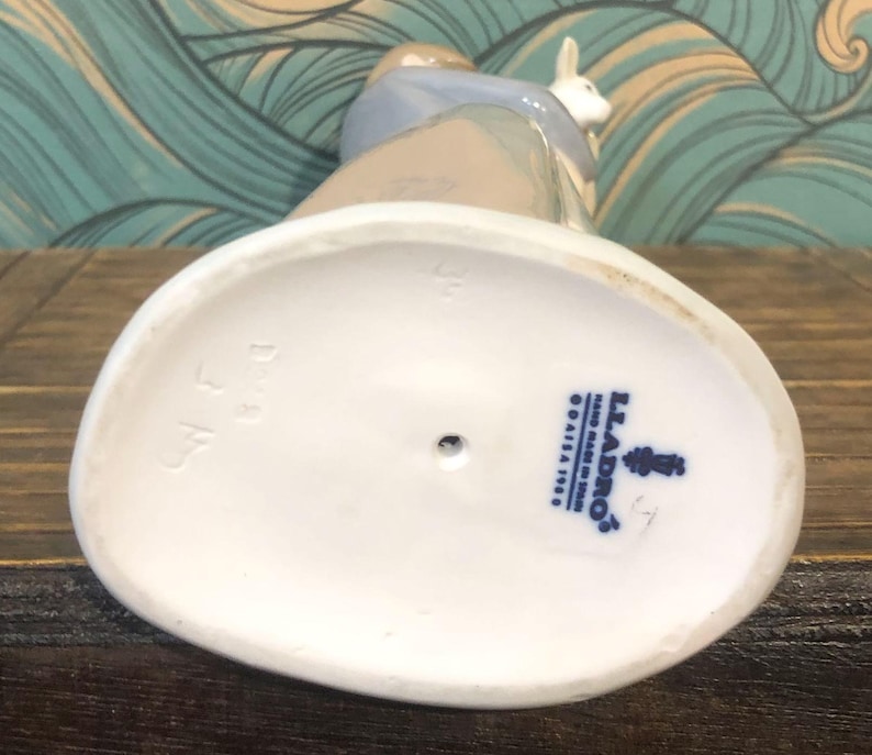 May include: A porcelain figurine of a person with a rabbit. The figurine is primarily white, with accents of blue, tan, and brown. The base is oval-shaped and white, with the Lladro logo and text. The figurine is on a wooden surface.