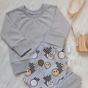 Könnte beinhalten: Ein hellgraues Langarm-Sweatshirt mit passender Hose, die ein sich wiederholendes Muster aus gelben Smileys, schwarz-weißen Karomustern und weißen Peace-Zeichen auf hellgrauem Hintergrund aufweist.